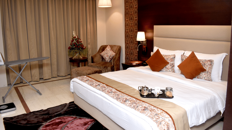 A bedroom in Lords Plaza Surat with a double bed, plush pillows, patterned runner, large window, ironing table, and comfortable seating area