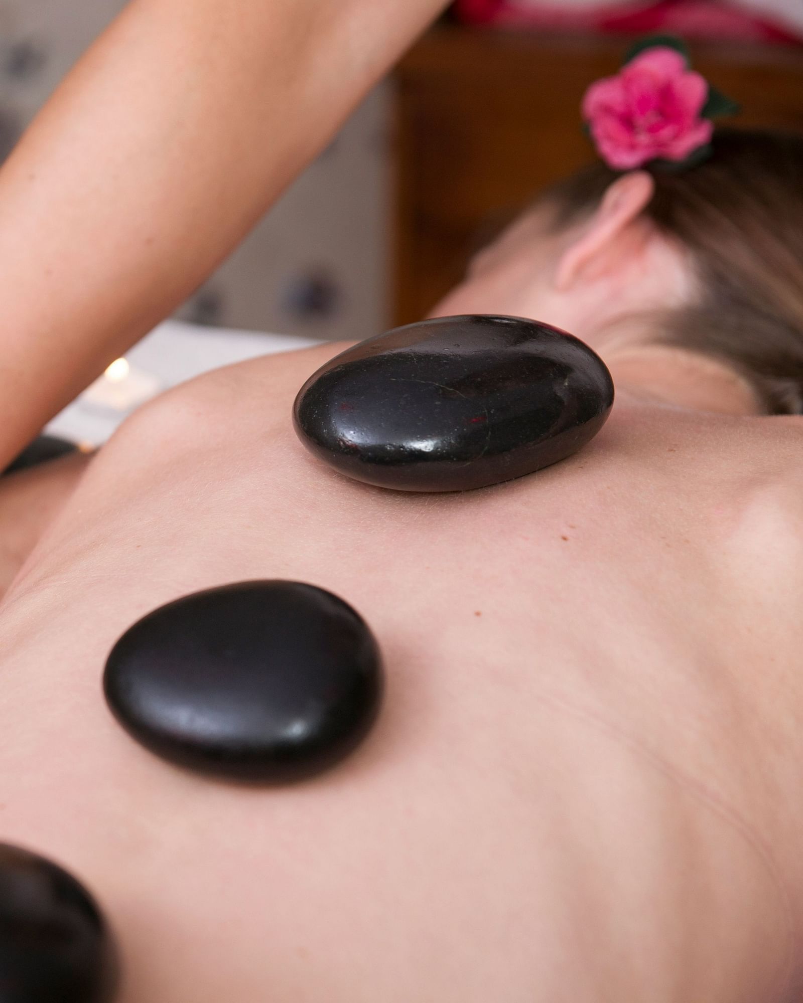 A person receiving a hot stone massage on their back, with smooth stones placed along their spine and a therapist's hands working on their shoulder. A flower adorns the person's hair.