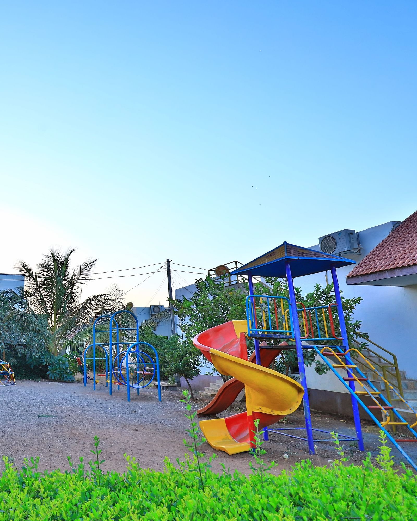 A vibrant outdoor children's play area with swings and slides, surrounded by green grass and palm trees - Vishal Lords Inn Gir Forest