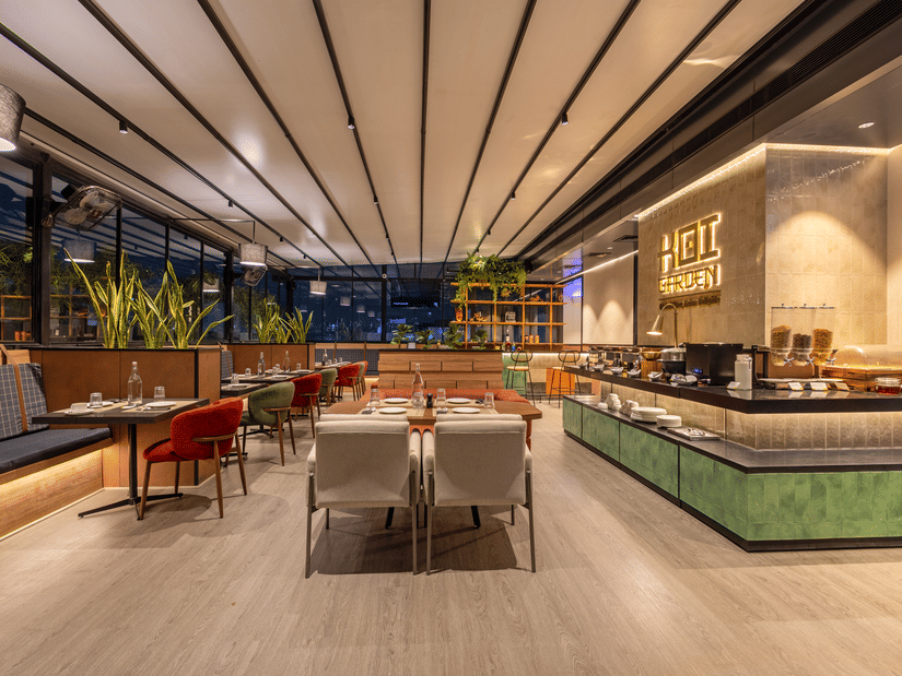 An expansive view of the Koi Garden dining room at Hotel Bawa Gurgaon during the evening. Multiple tables are prepared for guests across the floor. A buffet station is positioned along the right side.
