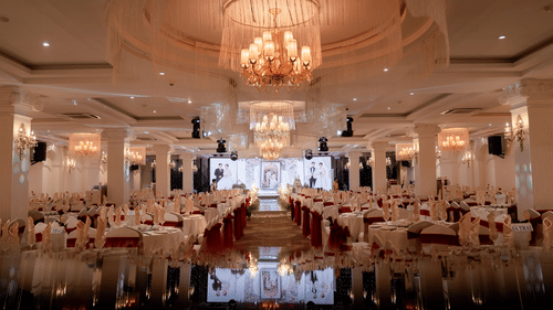 A grand banquet ballroom with ornate columns and crystal chandeliers, rows of round tables in white and burgundy linen, set for a wedding reception.