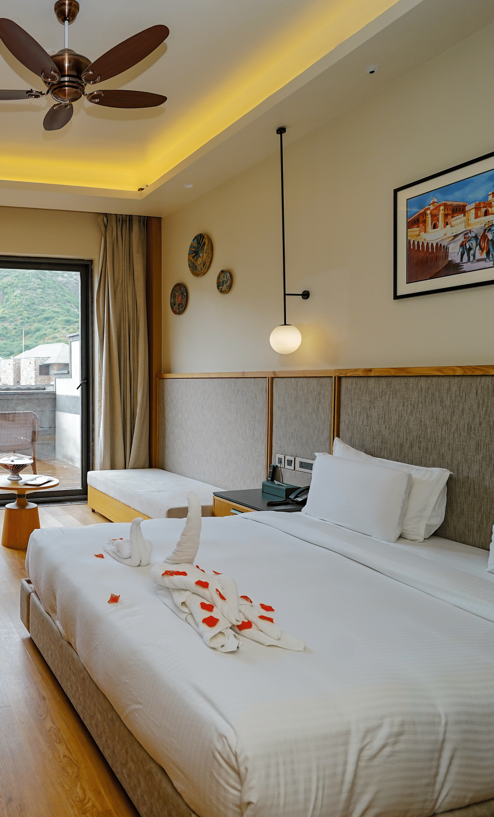 A well-lit hotel room with a large bed, a television, and a sliding glass door leading to a patio. There are rose petals on the bed.