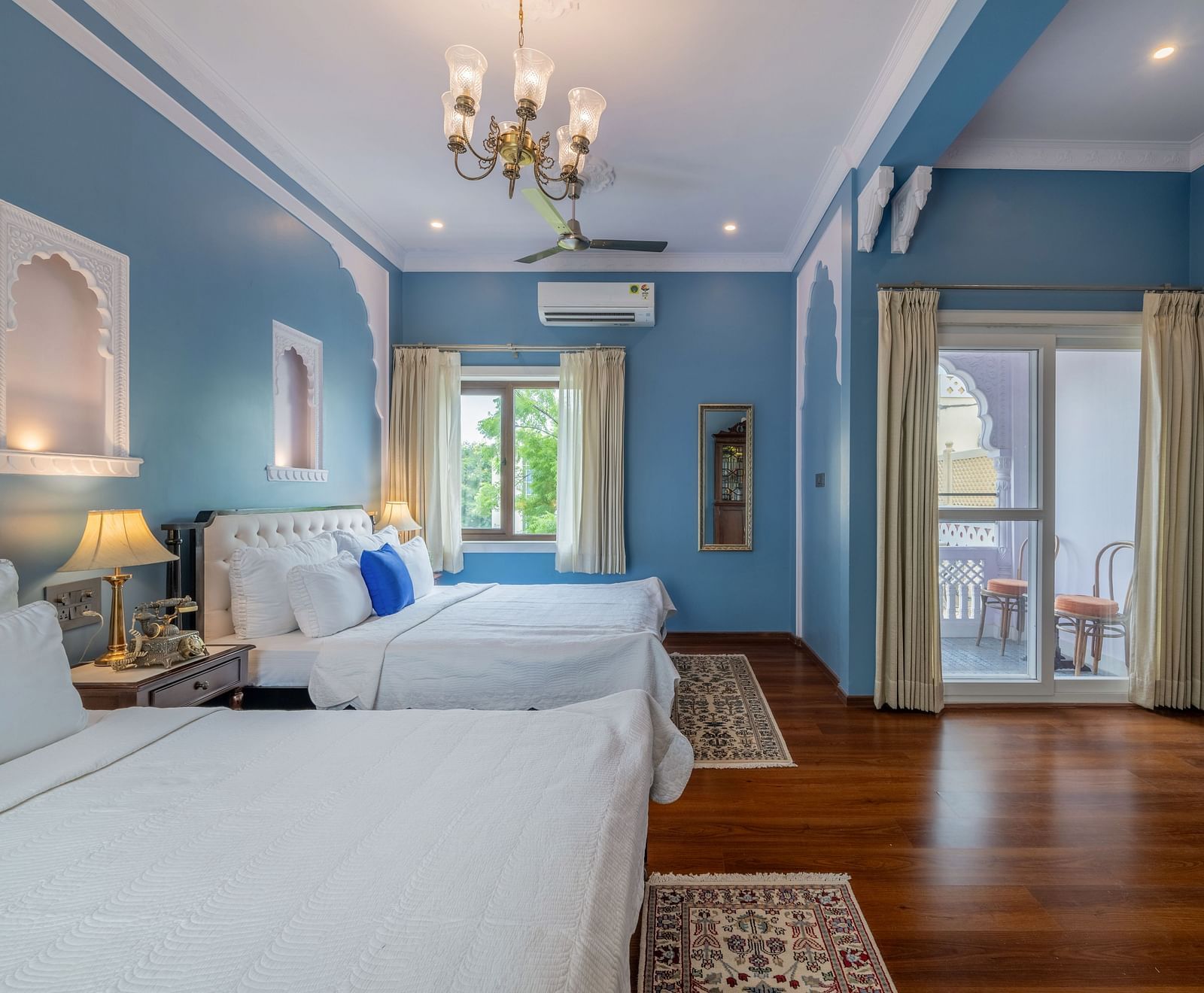 A bright and airy room at Dera Rawatsar, Jaipur, offers two comfortable beds, a balcony with seating, elegant wall details, and wooden flooring.