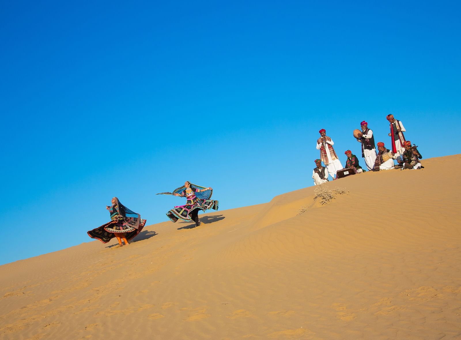  A cultural scene unfolds on a desert dune, with individuals dressed in traditional attire, bringing life and color to the sandy environment.
