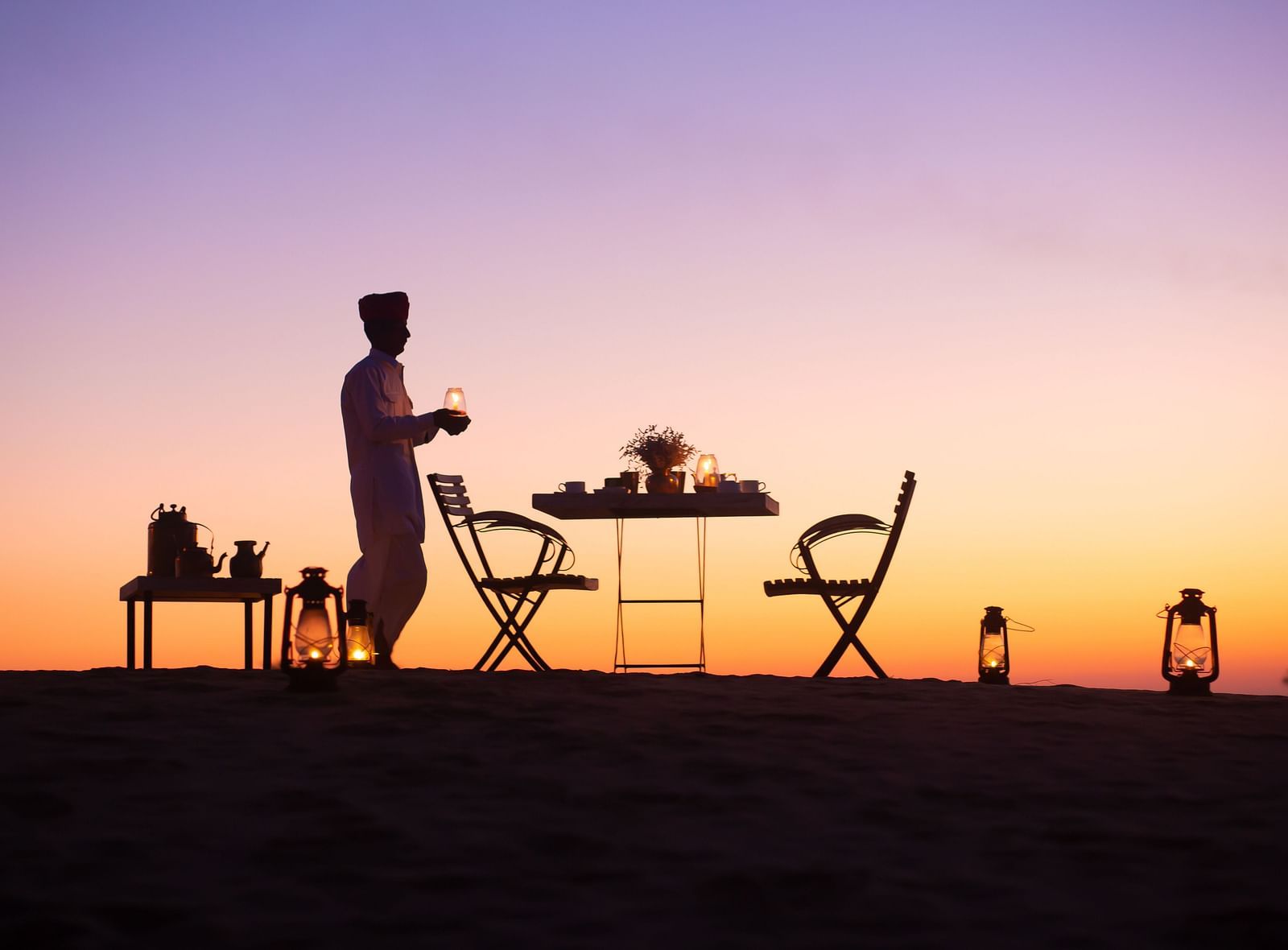 An evening dining setup in the desert features a table for two, illuminated by the last light of day and surrounded by softly glowing lanterns