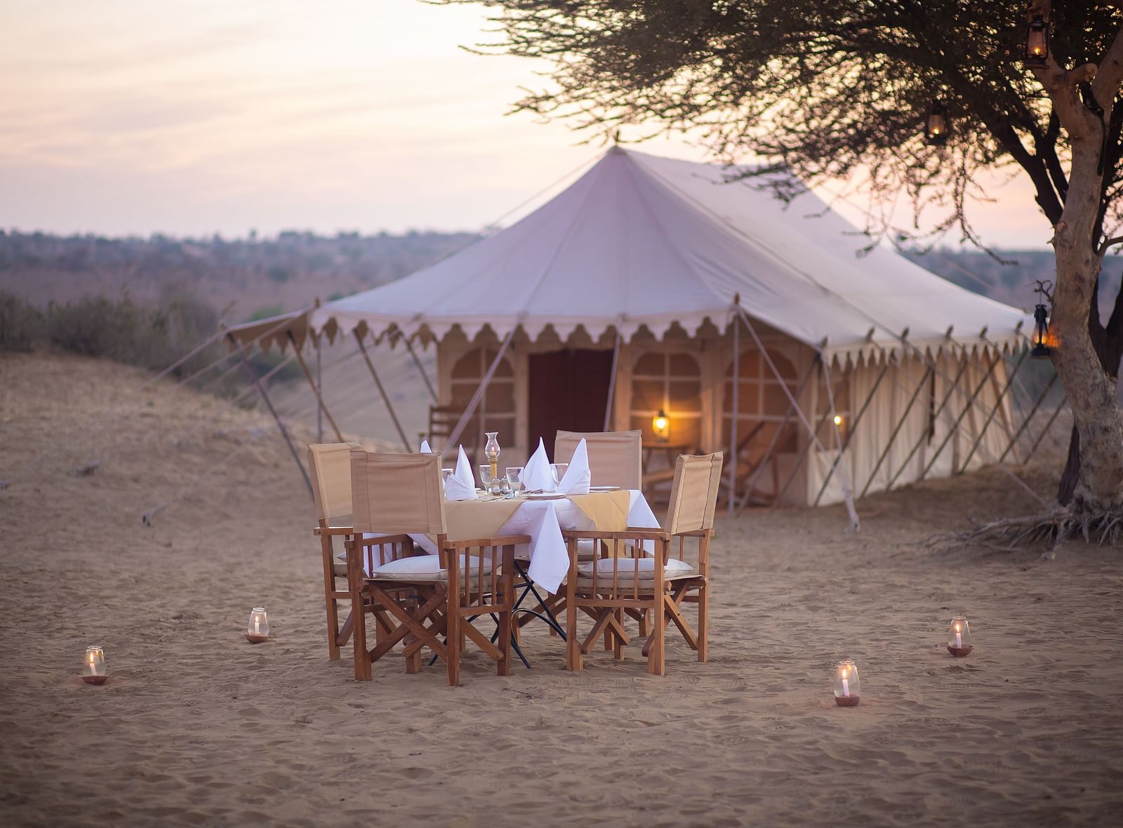 An outdoor dining set awaits by a large tent in the desert, shaded by a tree, for an intimate meal amidst the tranquility of nature.- at Manvar Resort 