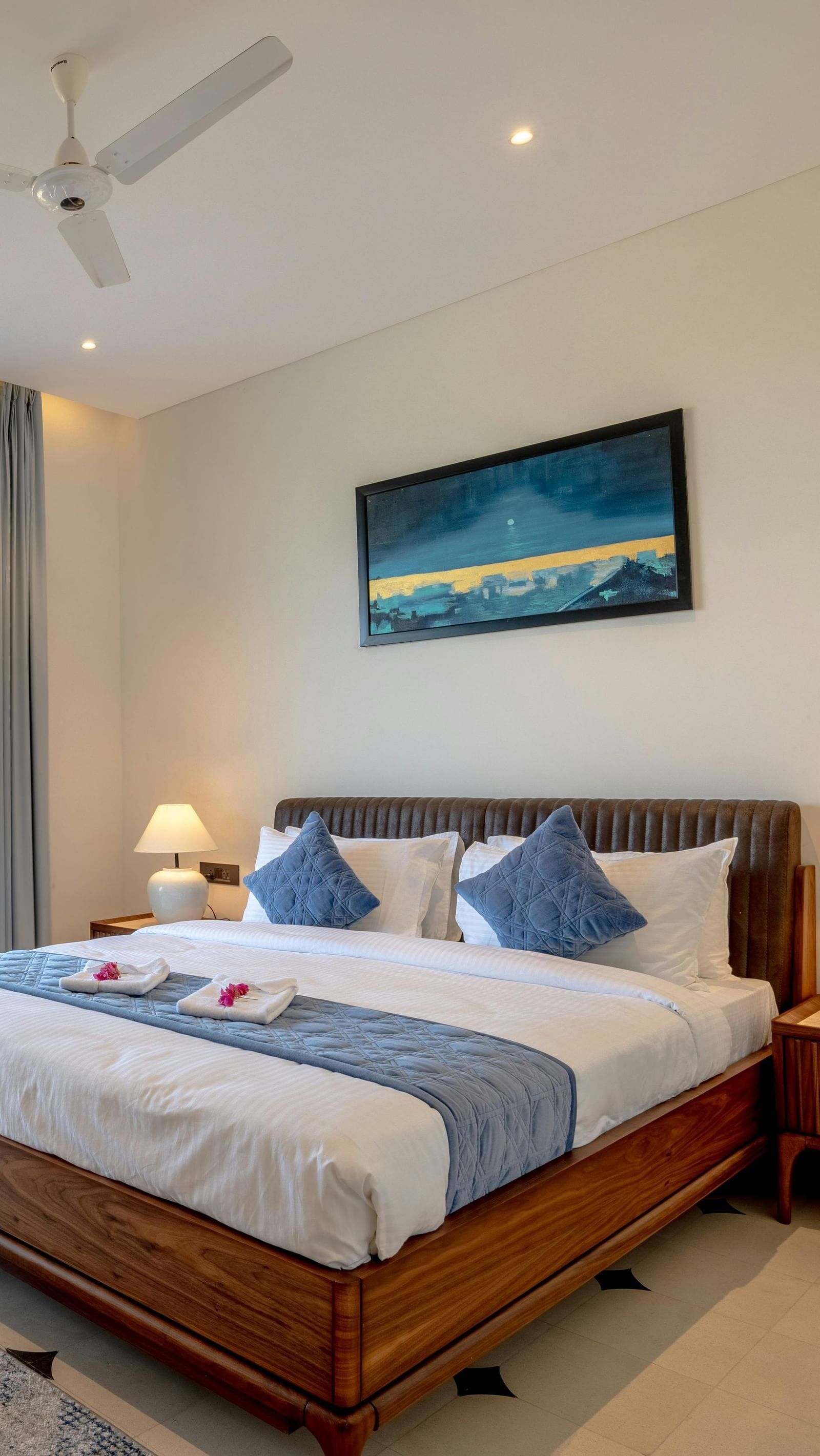 A room with balcony, spacious bed with bedside tables and lamps, artwork above the headboard, and outdoor view through glass doors