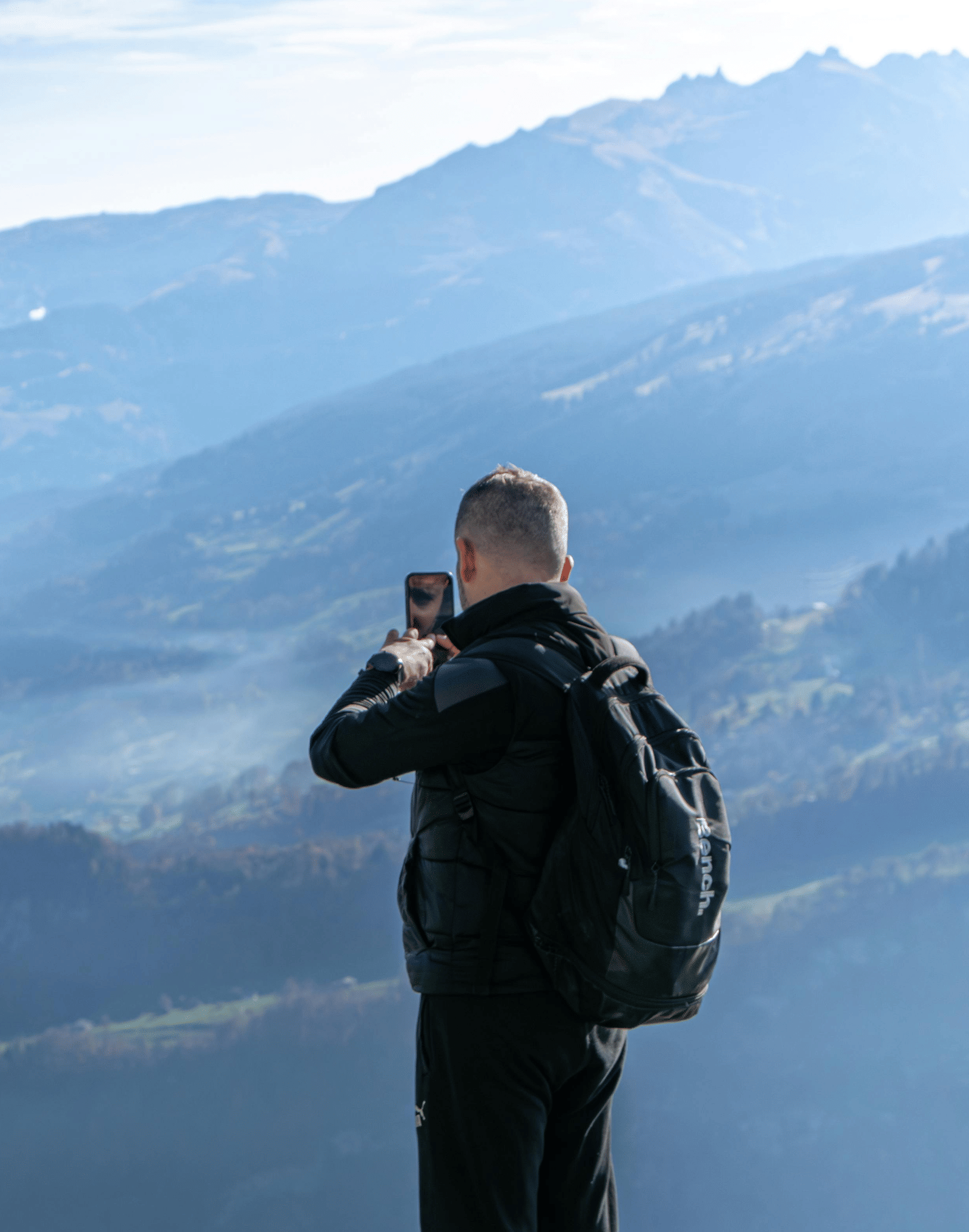 A man standing on a view-point clicking pictures of the valley and mountains spread in front of him.