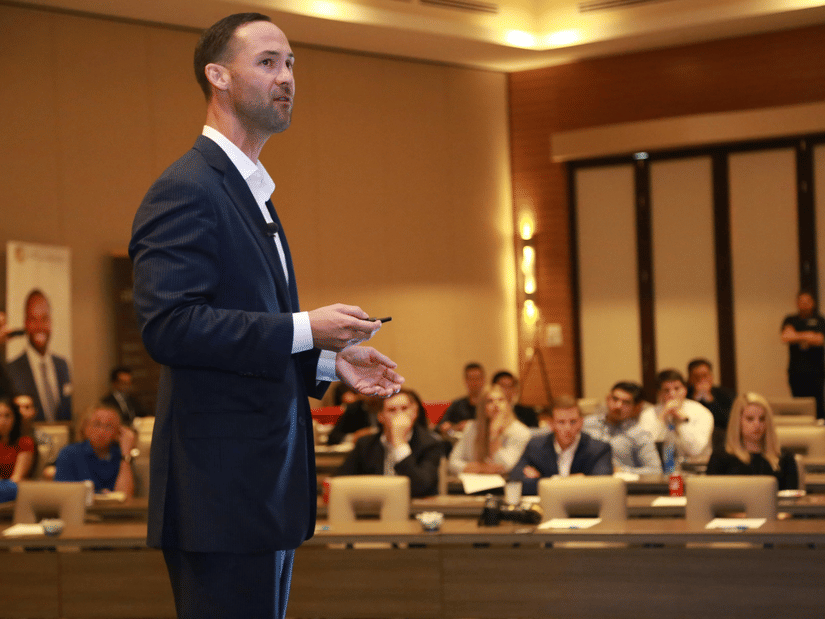 A man in a suit is giving a presentation or speech to an audience seated at tables.