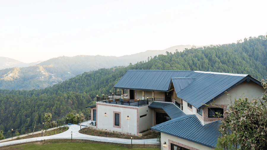 The buildings seen from above at dusk, with pitched metal roofs, a railed upper terrace, lit wall lamps and a paved drive at The Chamomile Estate.