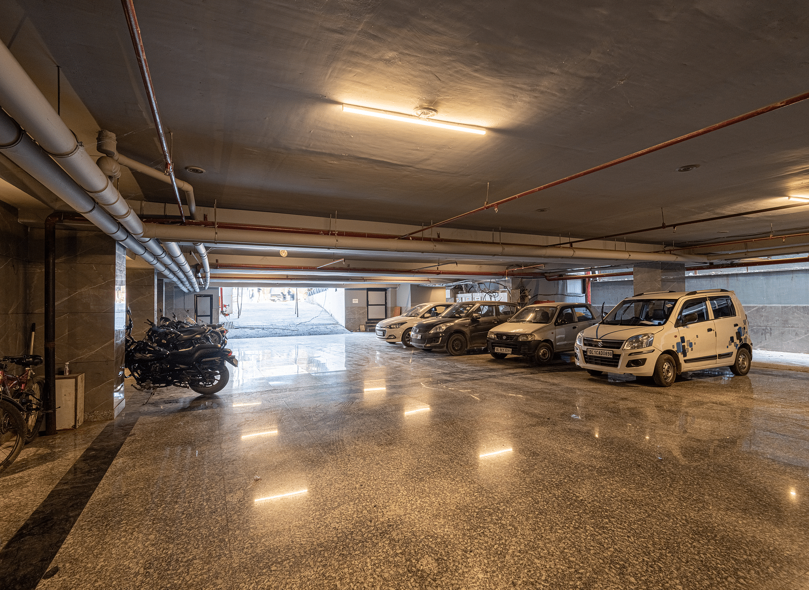 Image of a large parking space at DLS Grand Luxotica, Dehradun featuring cars and bikes parked in warm lighting
