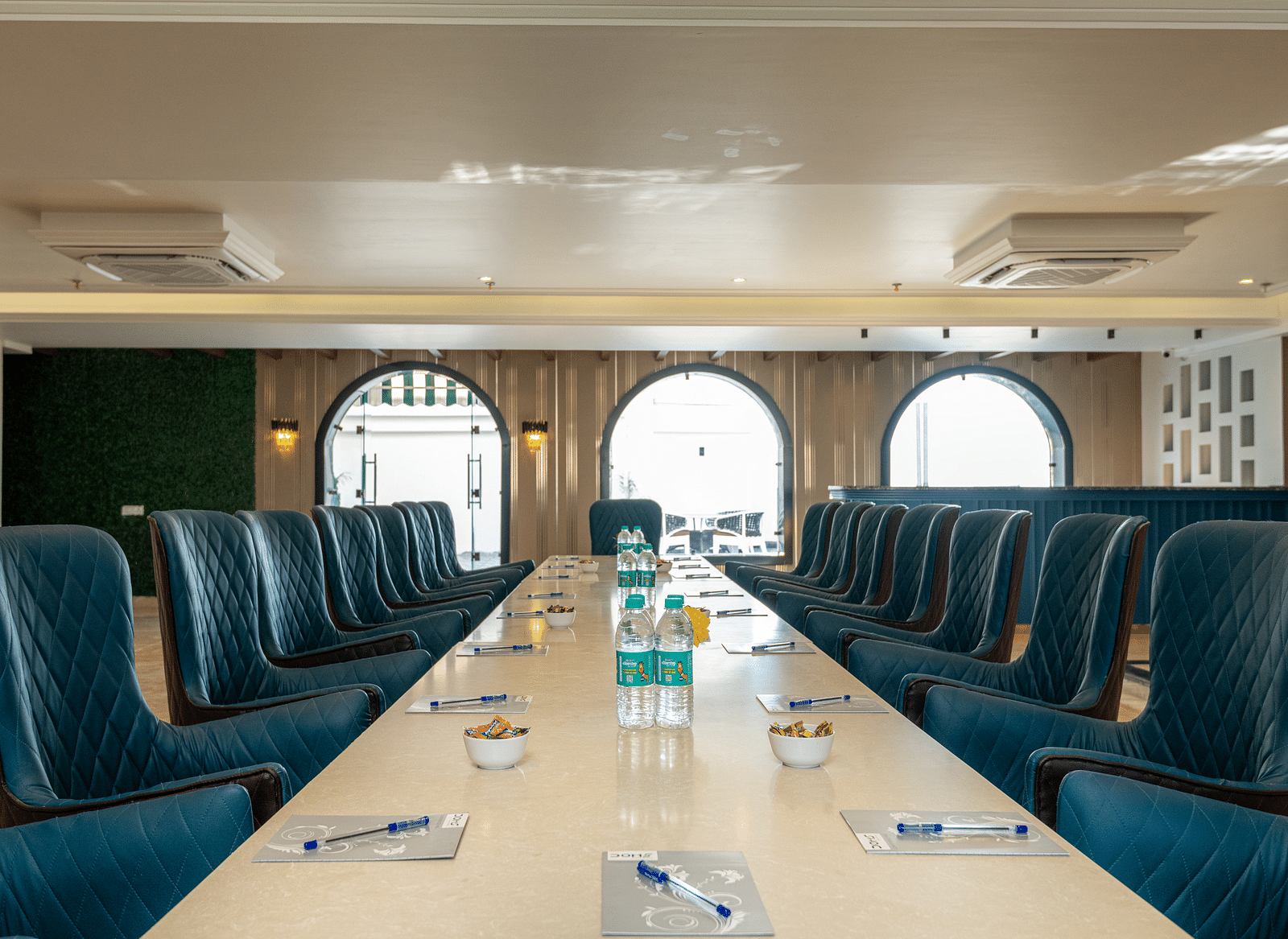 chairs arranged with a large table with notepads and pens along with water bottles on it as the daylight makes its way through the windows in the background