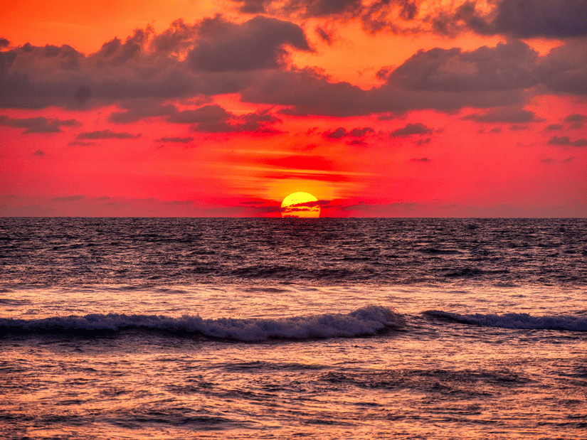 A vibrant sunset over the ocean, with the sun low on the horizon against a cloudy sky, reflecting deep orange and pink hues on the gentle waves.