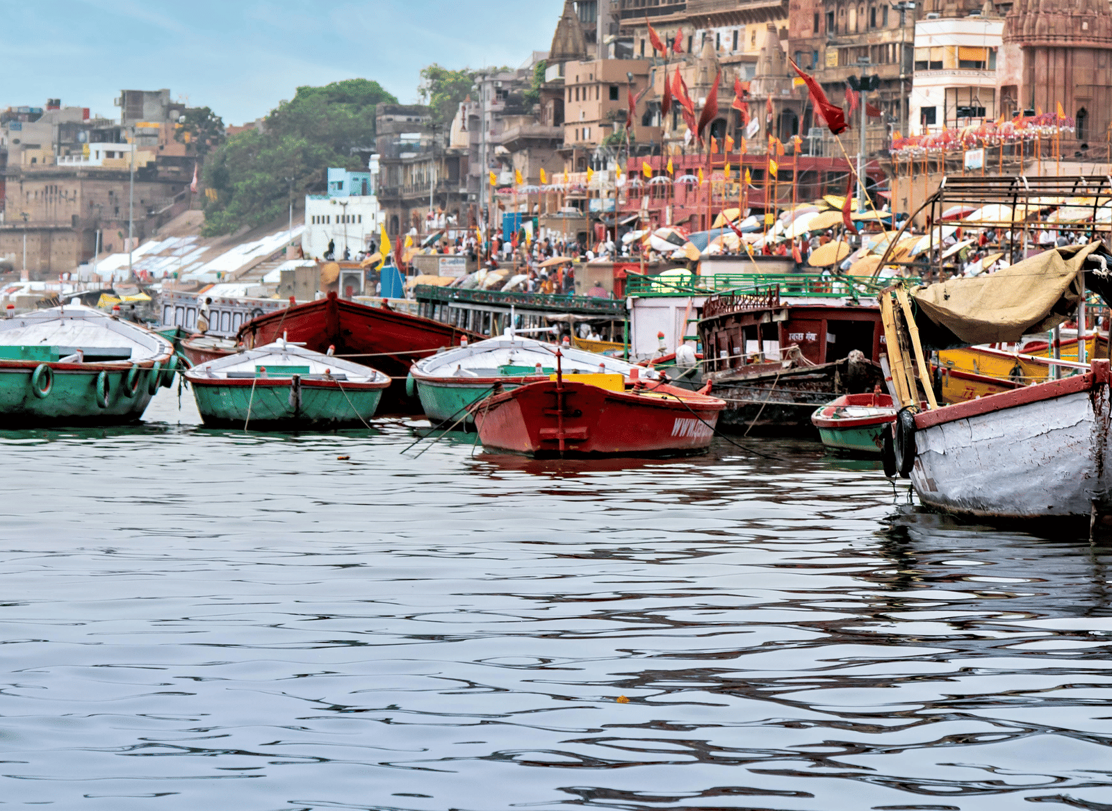 Small wooden boats moored on the Ganges River near the steps (ghats) of a crowded riverside city in India.