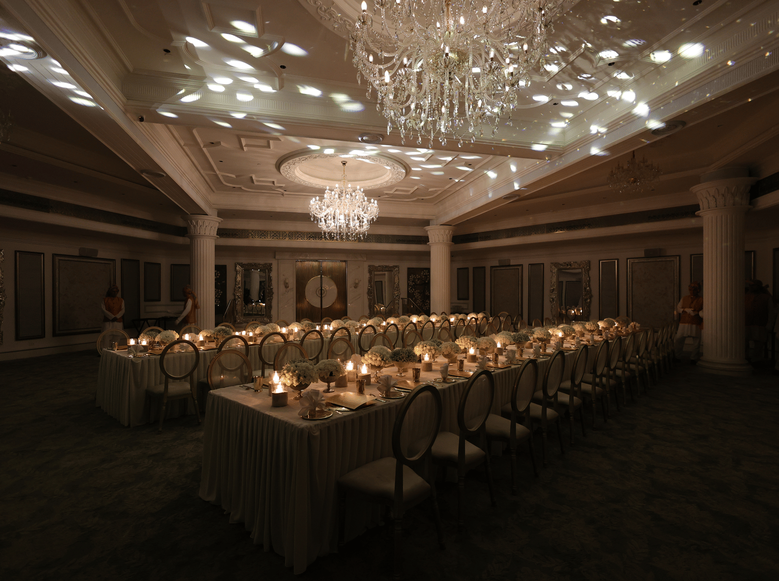 Dimly lit luxury banquet hall featuring a long dining table setup with candles and decorative ceiling light projections under chandeliers.
