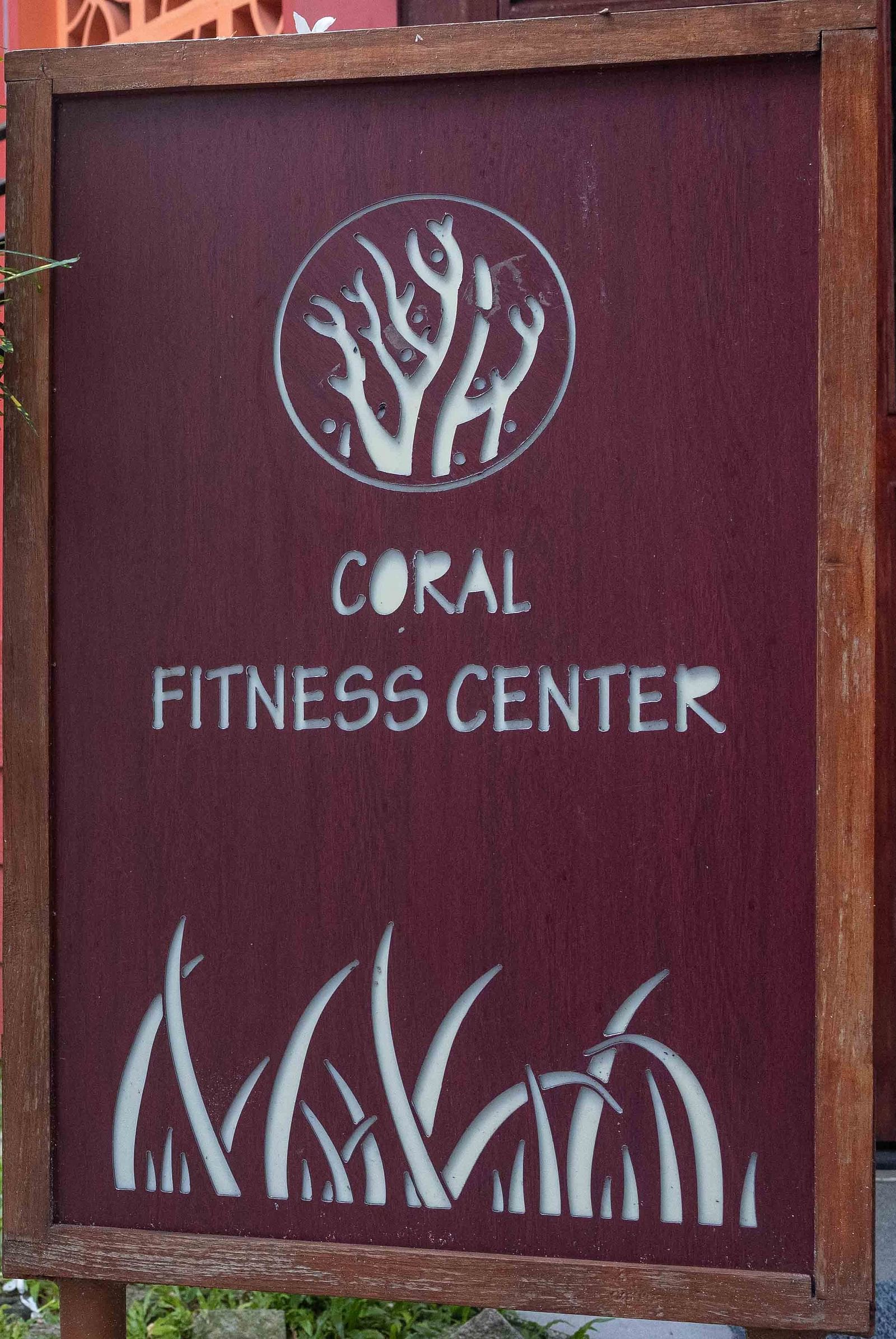 A sign for the Coral Fitness Center featuring sleek, modern design elements and a tree logo. The sign is set against a vibrant green garden, conveying a healthy and refreshing vibe - Coral Reef Resort & Spa, Havelock