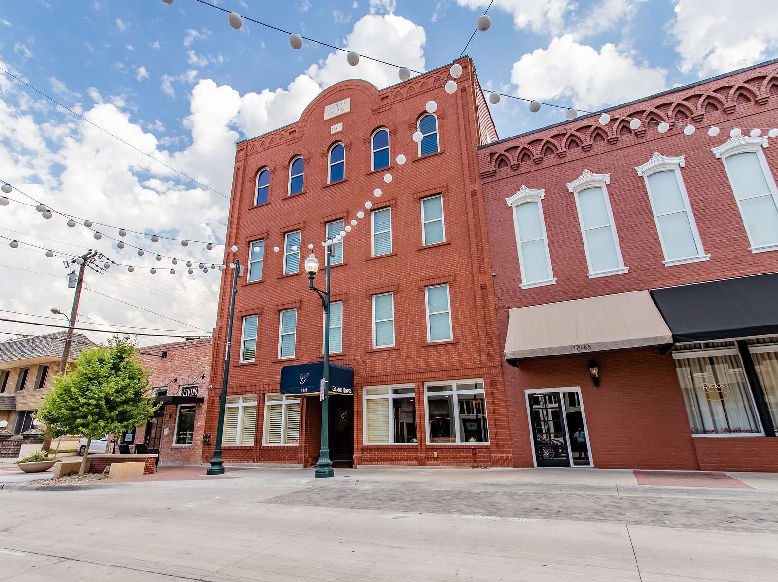 A multi-story brick building with large windows, awnings, and string lights hanging above the street.- The Grand Hotel & Ballroom, McKinney