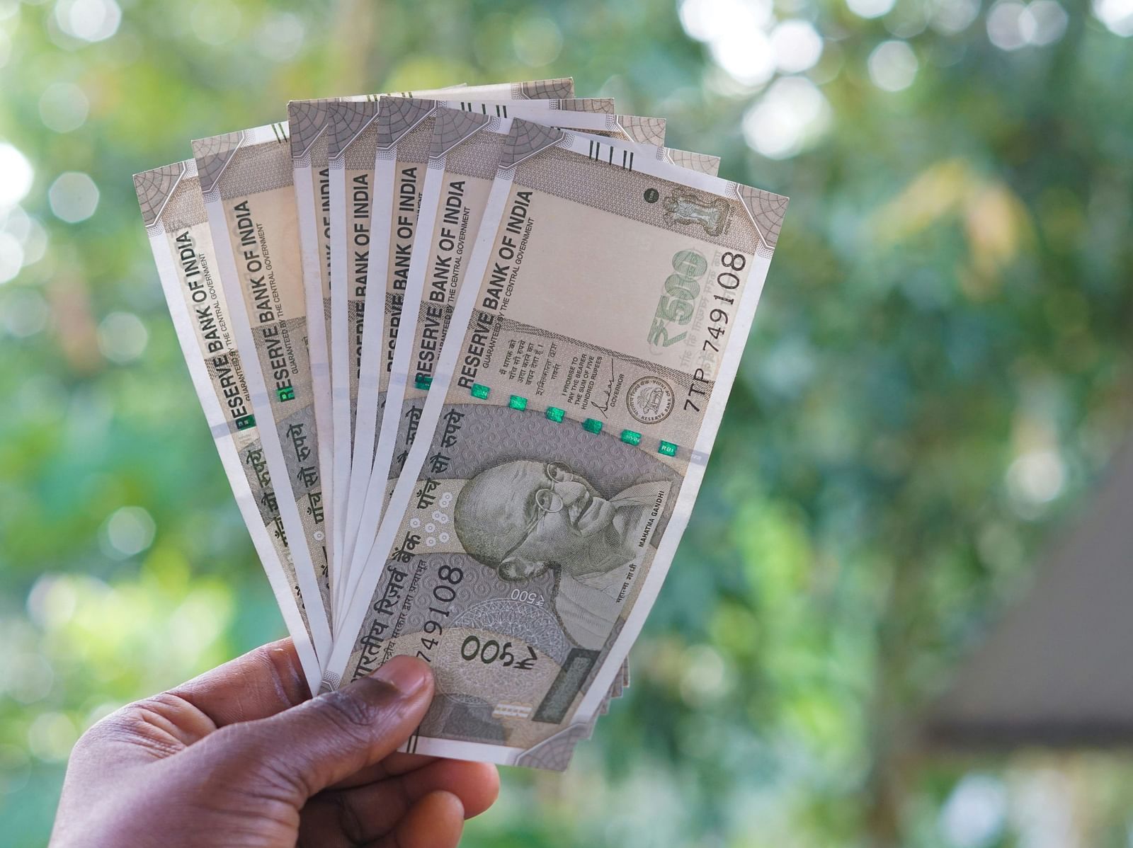 A close up of a person holding seven notes of 500 rupees with the background blurred.