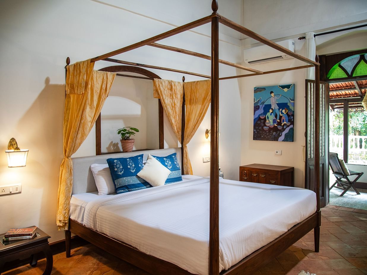 An image of four poster bed with yellow curtains,bedside table with a lamp, a cabinet at one corner, and an attached balcony where a chair is place - Lamrin Ucassaim Hotel, Goa