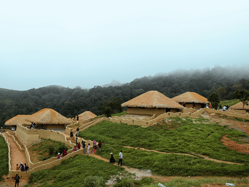 Earthen huts with thatched roofs built into a grassy hillside in En Ooru tribal village Wayanad, Kerala, with people walking on paths and mist covering the forest behind.