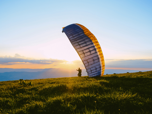 Colourful paraglider preparing for take-off across grassy hills, silhouetted against a warm sunrise with expansive open countryside beyond