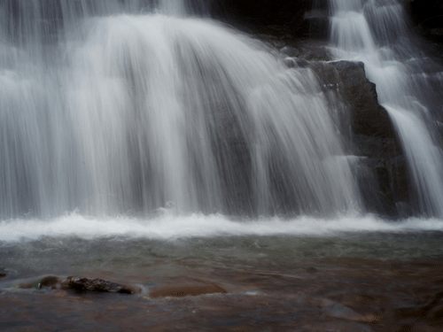 A smooth-flowing waterfall captured in motion, with water tumbling over dark rocks into a shallow pool.