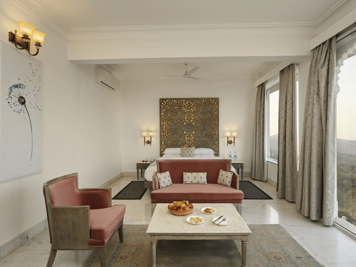 Seating area of Plunge Pool Suite in Fateh Safari Suites featuring sofa sets, a centre table with fruit basket, cookies, and other items, bedside tables on either side, and a huge window with view of greenery.