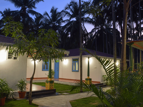 A white resort cottage with a red roof and blue window frames, surrounded by palm trees and green lawns - Ibex Resorts, Coimbatore (Kakarla)