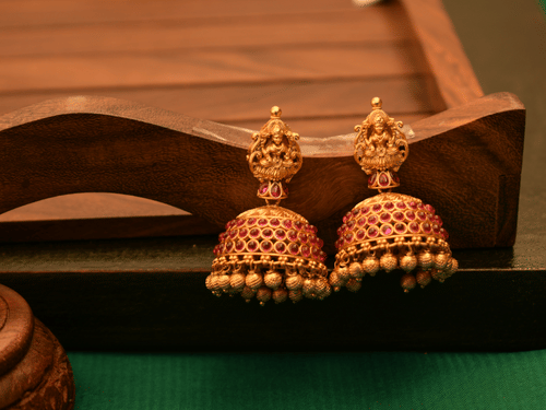 A pair of traditional Indian gold jhumka earrings, intricately studded with rubies, rests on a dark wooden tray.