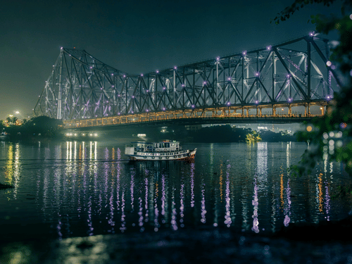 Howrah Bridge illuminated at night, reflecting on calm river waters, with boats drifting beneath and city lights shimmering softly