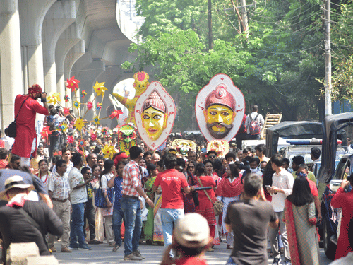 A procession on a road with many people carrying busts and some taking pictures on a pole next to parked cars during the Bengali New Year in Kolkata