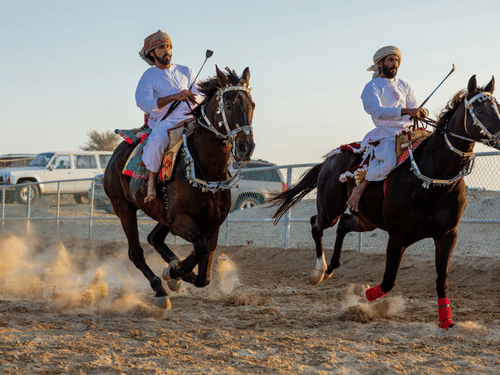 2 men in traditional white robes and turbans gallop dark horses across a dusty desert track at high speed.