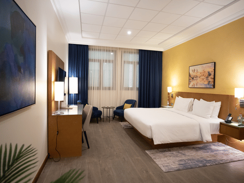 Spacious bedroom with large bed with white linens, two stools, yellow feature wall, and windows with blue and sheer curtains.