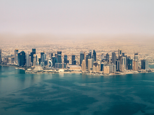 An aerial view of the Doha, Qatar skyline featuring modern skyscrapers along the blue Persian Gulf coast under a hazy sky.