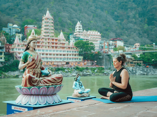 A woman practicing yoga in a seated pose by a river, with large Hindu statues and a multi-storey temple behind her.