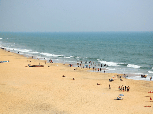 A high-angle view of a wide, golden sand beach meeting the turquoise ocean, with small groups of people and a few boats scattered along the shore.