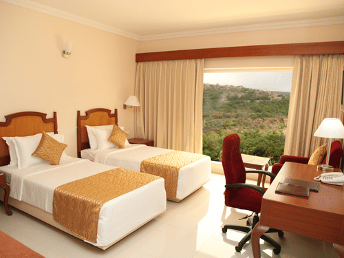 One of Raj Park Hotel's superior rooms in Tirupati featuring twin beds with gold runners, a wooden desk with a red chair, and a large window overlooking a forested hillside.