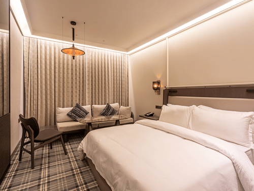 Modern hotel room at The Residency Towers Chennai featuring a large bed, seating area with sofa and chair, patterned carpet, and warm ambient lighting.