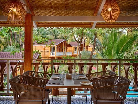 View from a covered dining area with woven chairs, a set dining table, overlooking wooden cabins among palm trees Aralea Beach Resort by Stone Wood, Morjim