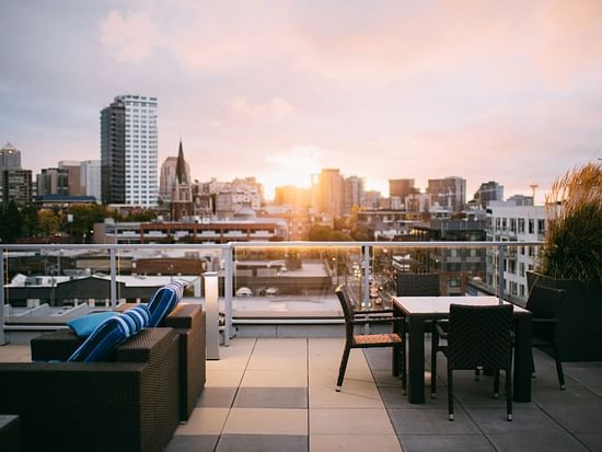 Rooftop terrace with tables, chairs, and sofas overlooking the city skyline during sunset.
