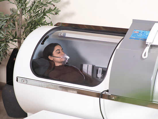 A person lying down inside a white hyperbaric oxygen chamber with a transparent window, wearing a specialized breathing mask.