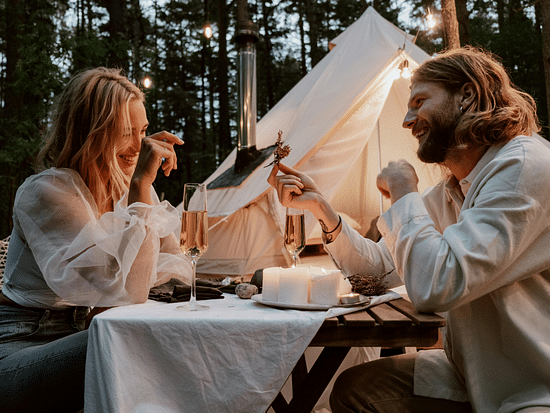 A couple laughing and toasting with champagne at an intimate candlelit outdoor dinner in a forest setting
