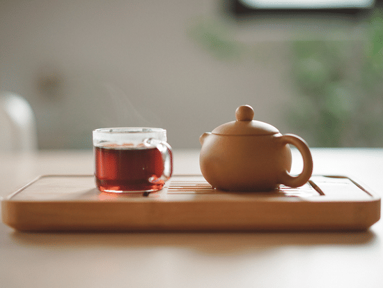 an image of a glass tea cup kept next to an earthen kettle on a tray inside a room