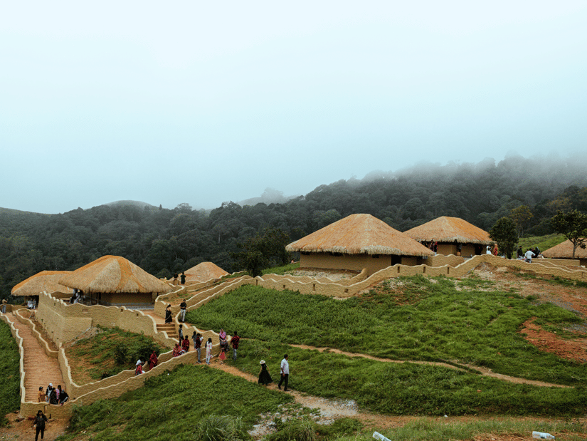 Earthen huts with thatched roofs built into a grassy hillside in En Ooru tribal village Wayanad, Kerala, with people walking on paths and mist covering the forest behind.