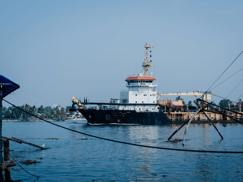 A large industrial ship glides calmly through coastal harbour waters near fishing nets suspended along the shore.