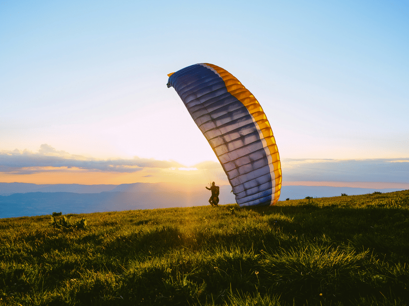 Colourful paraglider preparing for take-off across grassy hills, silhouetted against a warm sunrise with expansive open countryside beyond