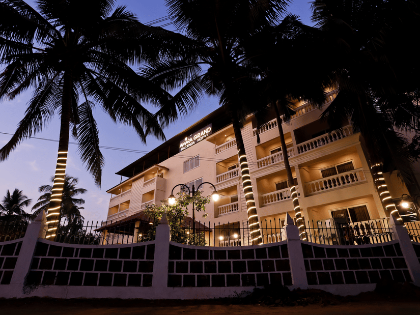 Exterior view of Amara Grand Baga, Goa, with a multi-storey building and trees on the side under a blue sky.