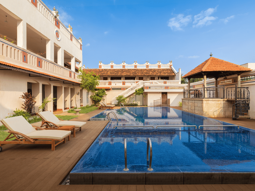 An outdoor swimming pool at Chidambara Vilas, a Chettinad heritage stay, surrounded by heritage-style architecture with lounge chairs and a shaded pavilion overlooking the courtyard