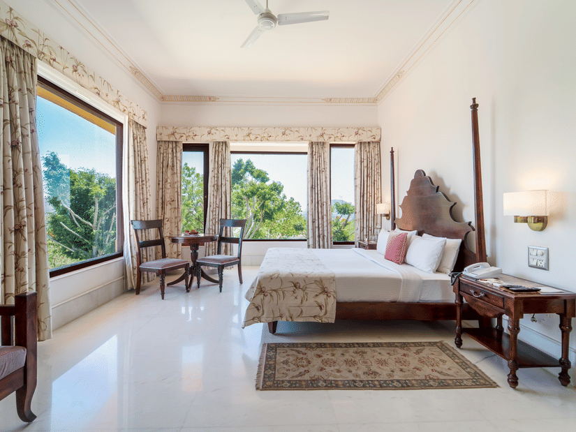 A spacious bedroom featuring a dark wood 2-poster bed, elegant drapery, and large windows with garden views - Fateh Garh, Udaipur