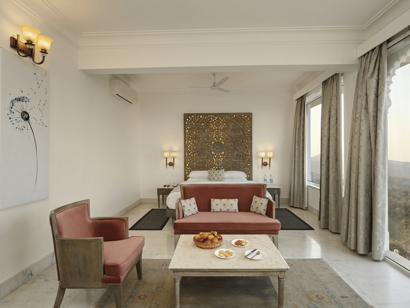 Seating area of Plunge Pool Suite in Fateh Safari Suites featuring sofa sets, a centre table with fruit basket, cookies, and other items, bedside tables on either side, and a huge window with view of greenery.
